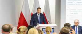 Minister Czarnek przy mównicy, za nim falgi biało-czerwone, na pierwszym planie duża grupa osób siedzących plecami do obiektywu 