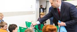 Minister nachyla się nad rzędem krzeseł, na których siedzą dzieci, i podaje coś małemu chłopcu w szarej bluzie.