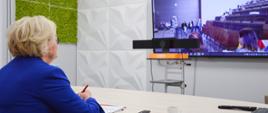 Wiceminister Machałek siedzi przy biurku, przed nią duży monitor na którym widać salę z ustawionymi w rzędy krzesłami.