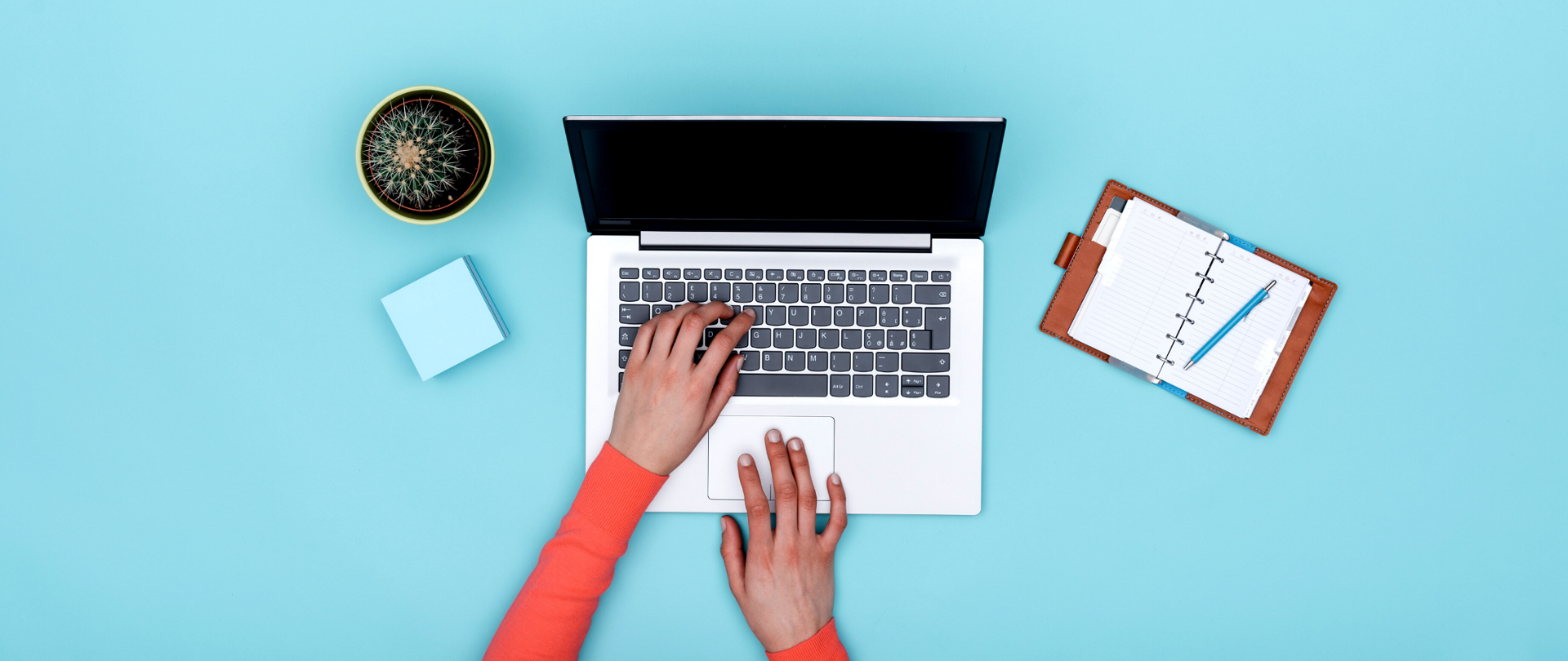Dłonie pracujące na otwartym laptopie. Obok laptopa leżą notes i kaktus.