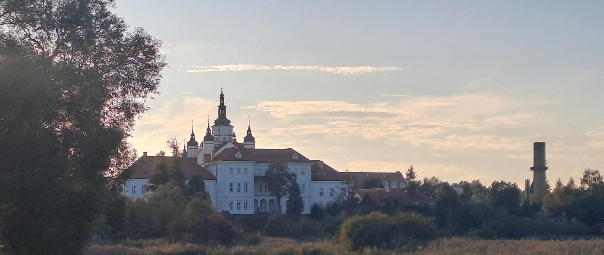 Biały zespół klasztorny z wieżami widziany z oddali ponad rzeką i terenami podmokłymi, odbijający się w spokojnej wodzie w świetle zachodzącego słońca.