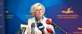 Wiceminister Machałek w białym ubraniu stoi na tle niebieskiej ścianki z napisem Karkonoska Akademia Nauk Stosowanych i mówi do kilku mikrofonów na stojakach.