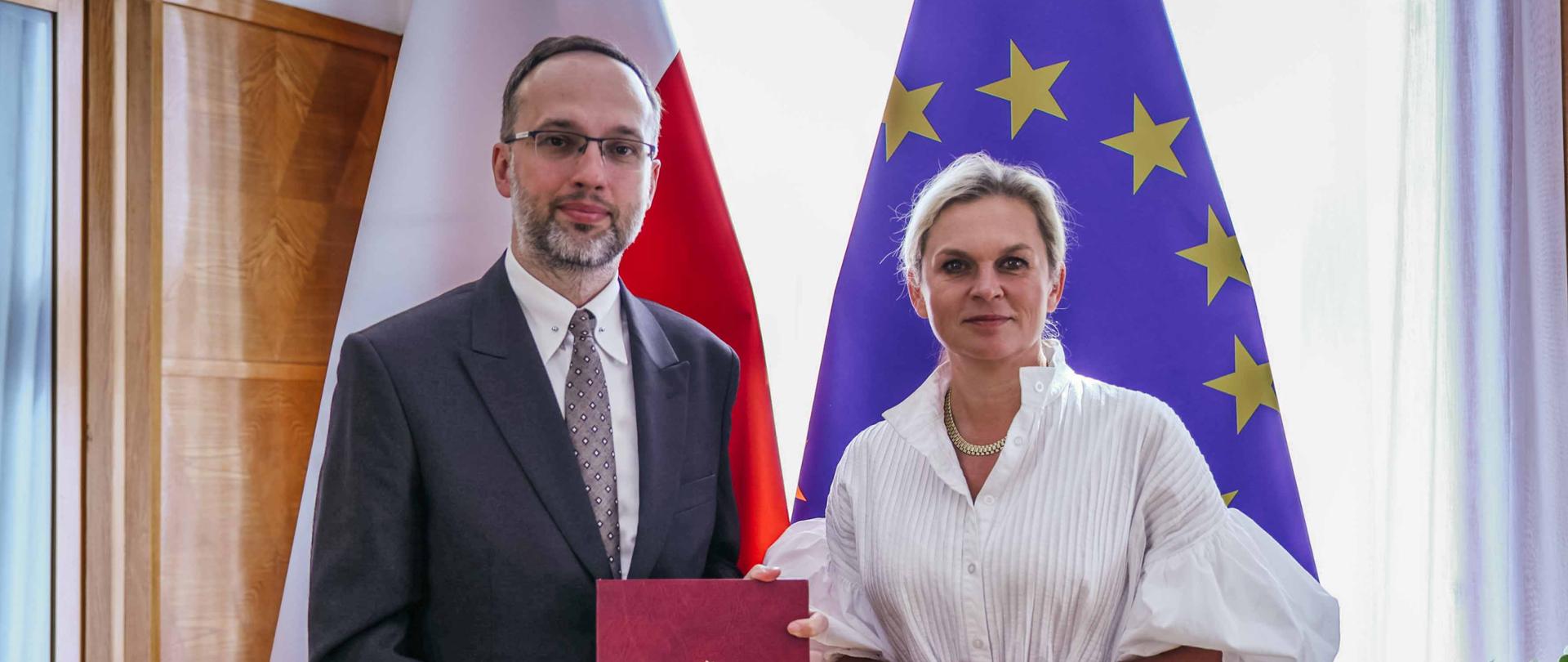 Mężczyzna i kobieta stoją obok siebie przed flagami Polski i Unii Europejskiej. Mężczyzna, w okularach i garniturze, trzyma w rękach czerwoną teczkę z polskim godłem. Kobieta, z włosami spiętymi w kok, ubrana jest w białą, luźną sukienkę z szerokimi rękawami, a w pasie ma zawiązany pasek. Na lewym nadgarstku ma sportowy zegarek. Oboje patrzą wprost na kamerę z poważnymi minami. Oświetlenie jest jasne, a tło sugeruje formalne spotkanie, prawdopodobnie w biurze.