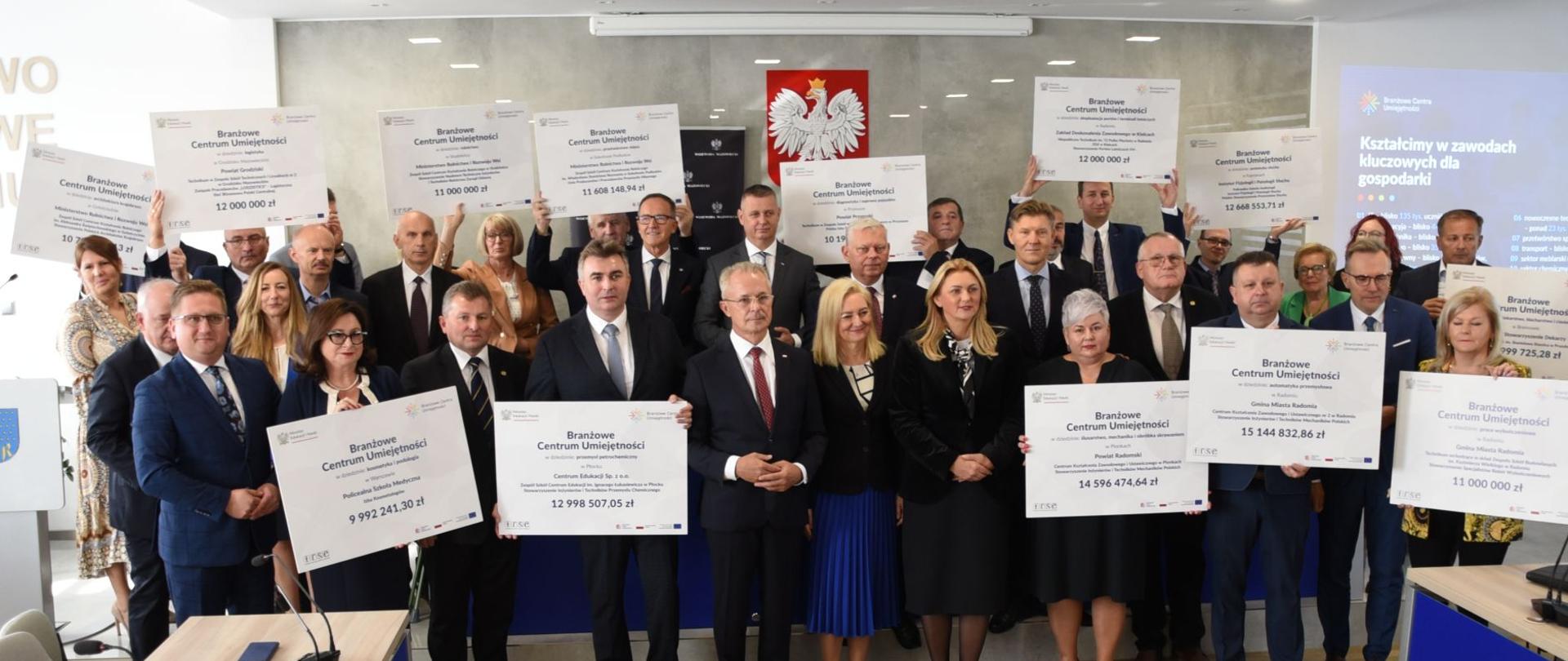 Zdjęcie grupowe, dużo osób stoi pod ścianą, wiele z nich trzyma duże karty z napisami Branżowe Centrum Umiejętności.