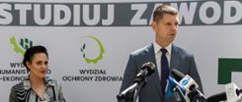 Minister Piontkowski stoi obok kolorowo ubranej kobiety i mówi do mikrofonu.