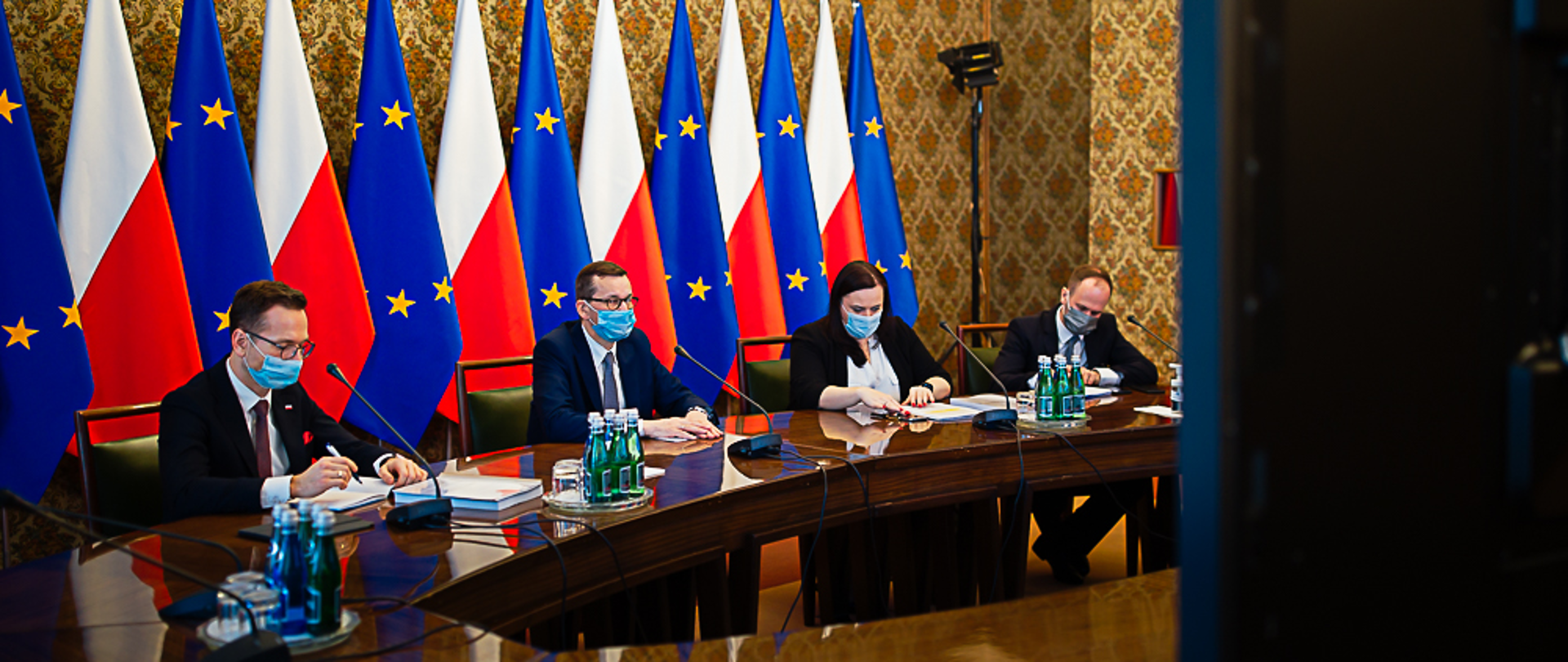 premier Mateusz Morawiecki, wiceministrowie Małgorzata Jarosińska-Jedynak i Waldemar Buda patrzą w kierunku uczestniczących online w spotkaniu przedsiębiorców, za ministrami flagi Polski i Unii Europejskiej 