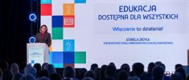 Kobieta (Podsekretarz Stanu Izabela Ziętka) w szarym garniturze stoi na mównicy i przemawia, z tytułem "EDUKACJA DOSTĘPNA DLA WSZYSTKICH. Włączanie to działanie!" oraz informacją o prelegentce. Scenę wypełniają duże, podświetlone kolorowe kwadraty z ikonami. Na pierwszym planie widoczna jest publiczność.