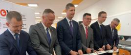 Sześciu uśmiechniętych mężczyzn w garniturach stoi w rzędzie, każdy trzyma nożyczki i przecina wiszącą w poprzek taśmę z napisami SOWA, za nimi duży baner z takim samym napisem.
