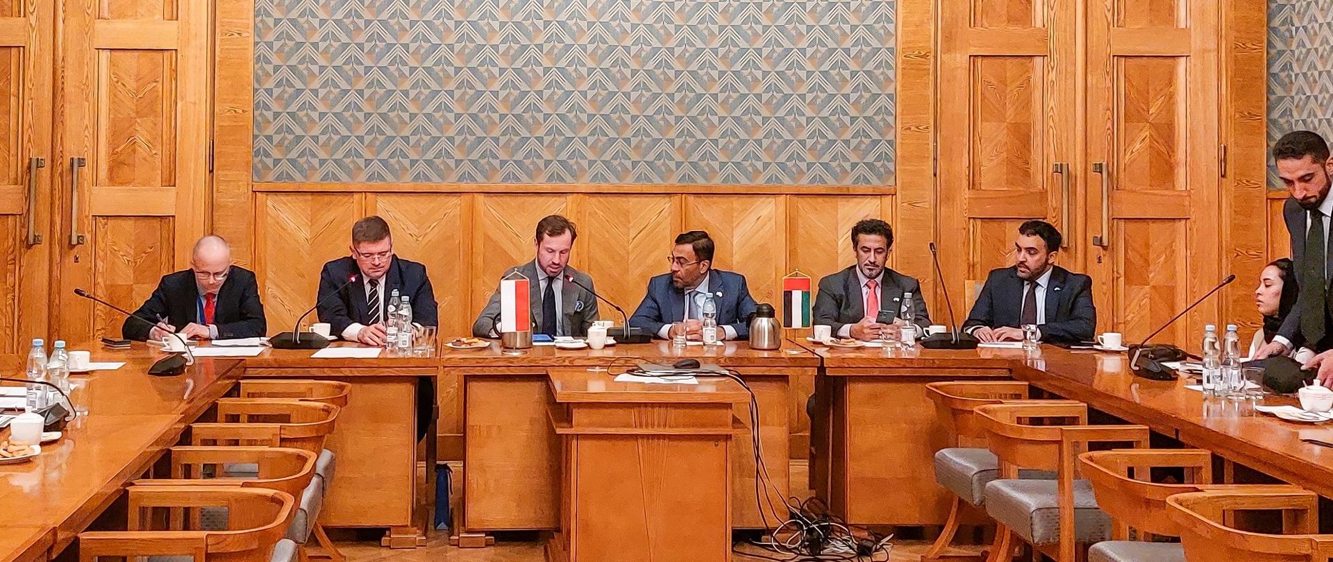 W dużej sali ze ścianami wyłożonymi drewnianymi panelami za drewnianym stołem siedzi grupa kilku osób.