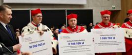 Kilku mężczyzn w czerwonych ceremonialnych strojach z gronostajowymi kołnierzami stoi w rzędzie, trzymają wielkie symboliczne czeki z napisami 10 000 000 zł, 2 746 180 zł, 1 998 700 zł, obok nich stoi minister Czarnek.