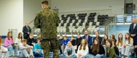 Scena z zajęć w hali sportowej. Żołnierz w mundurze polowym stoi tyłem, prowadząc prelekcję dla dużej grupy młodzieży siedzącej na niebieskiej podłodze, obok manekina do RKO. Minister Edukacji Barbara Nowacka i inni dorośli siedzą za młodzieżą.