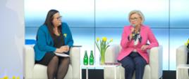 Przy małym białym stoliku na białym fotelu siedzi wiceminister Machałek w różowej marynarce, obok niej kobieta w niebieskiej marynarce , wiceminister mówi do trzymanego w ręku mikrofonu, na stoliku stoją żółte tulipany w wazonie.