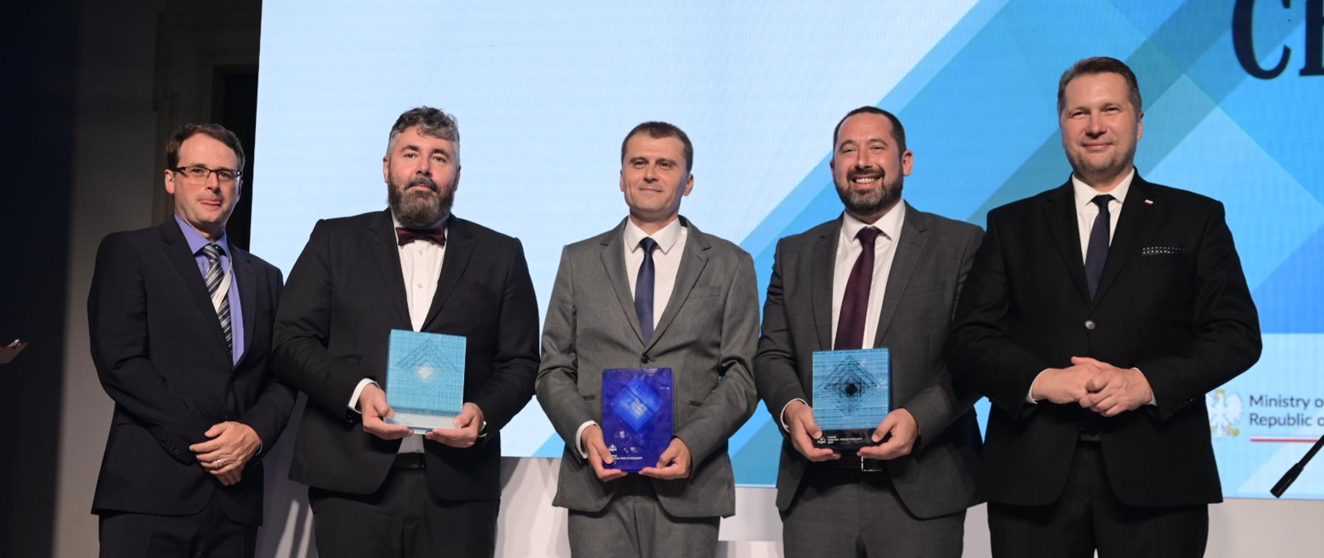 Pod wielkim ściennym ekranem stoi pięciu mężczyzn w garniturach, trzymają małe czworokątne niebieskie przedmioty.