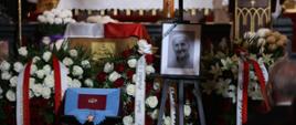 Widok na pokrytą biało-czerwonymi kwiatami trumnę, przed nią na czerwonej poduszce leży order w kształcie złotej gwiazdy, obok na stojaku czarno-białe zdjęcie.