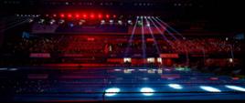 Widok na zaciemniony stadion, spod wysokiej konstrukcji na środek świeci kilka punktowych reflektorów.