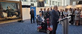 Widok na podwórko pomiędzy białymi budynkami, pod ścianą stoi obraz przedstawiający Kopernika, przed nim stoi wiceminister Murdzek i mówi do mikrofonu na stojaku, za nim mężczyzna na czterokołowym wózku inwalidzkim, w tle grupa ludzi.