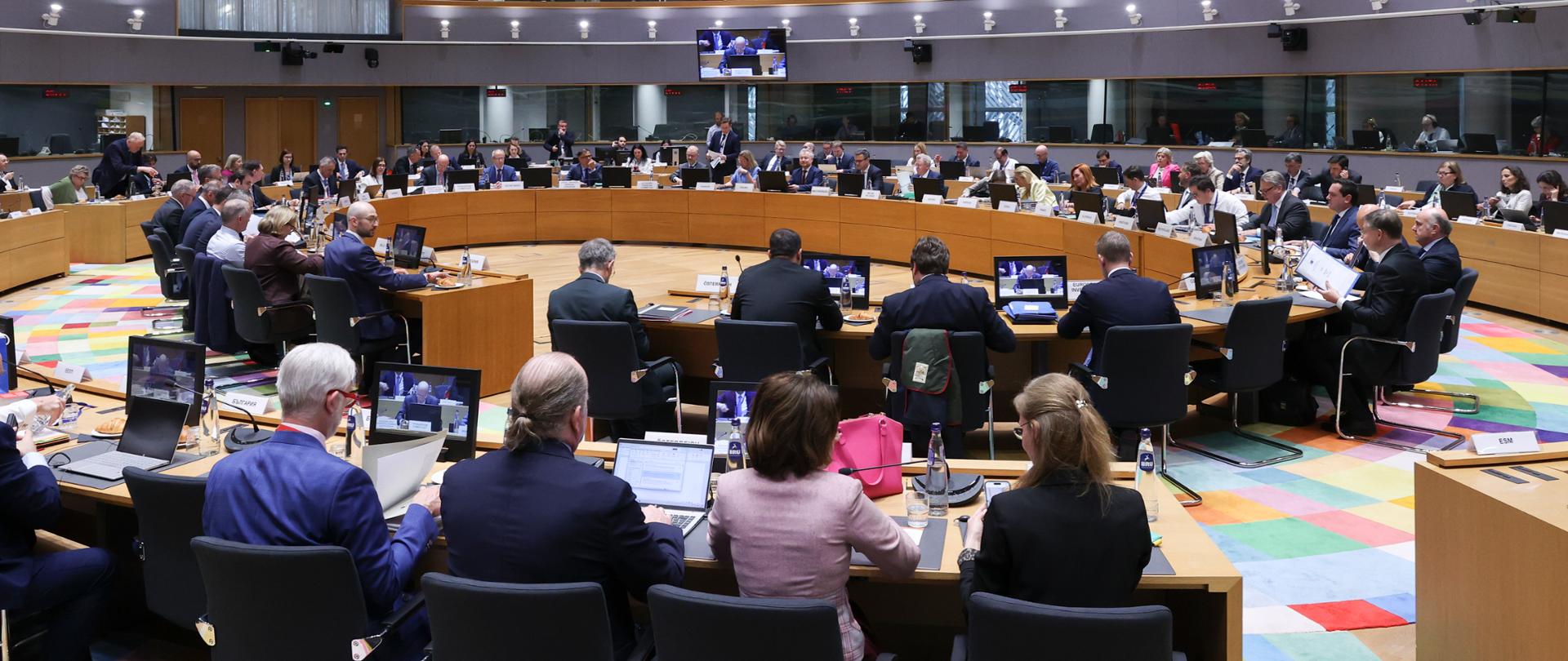 Sala obrad z uczestnikami posiedzenia ECOFIN przy okrągłym stole, pod kolorowym, geometrycznym sufitem z oświetleniem 