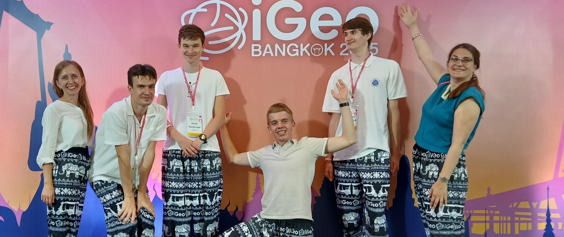 Sześcioro młodych ludzi stoi na scenie przed różowym tłem z napisem "iGeo BANGKOK 25". Wszyscy mają na sobie białe koszulki i ciemne spodnie z wzorem w słonie. Mężczyzna pośrodku kuca z rozłożonymi rękami, uśmiechając się do kamery. Pozostałe osoby stoją w rzędzie, uśmiechają się i machają. Oświetlenie jest jasne, równomierne, a atmosfera radosna i swobodna.