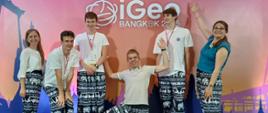 Sześcioro młodych ludzi stoi na scenie przed różowym tłem z napisem "iGeo BANGKOK 25". Wszyscy mają na sobie białe koszulki i ciemne spodnie z wzorem w słonie. Mężczyzna pośrodku kuca z rozłożonymi rękami, uśmiechając się do kamery. Pozostałe osoby stoją w rzędzie, uśmiechają się i machają. Oświetlenie jest jasne, równomierne, a atmosfera radosna i swobodna.