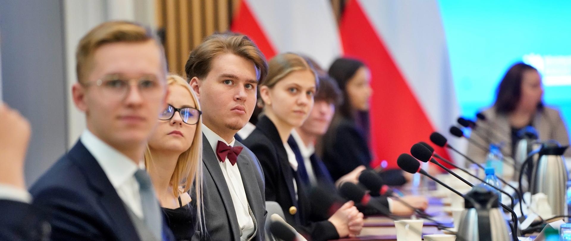 Widok z boku na lewą stronę sali, przy ustawionych w rząd stolikach siedzą młodzi ludzie, przed nimi mikrofony.