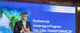 Kadr przedstawia zbliżenie na ekran i mężczyznę za mównicą, podczas konferencji otwierającej Program 'Zielona Transformacja'. Przemawia wiceminister Klimatu i Środowiska, Krzysztof Bolesta
