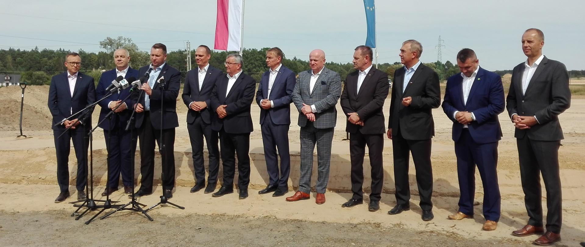 Uczestnicy uroczystości stoją w szeregu, przed nimi mikrofony
