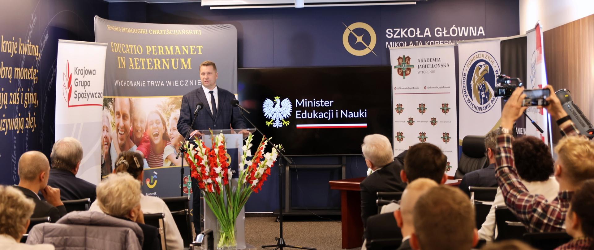 Widok z tyłu na wypełnioną siedzącymi na krzesłach ludźmi salę, przed nimi minister Czarnek stoi przy mównicy i mówi do mikrofonu, przed nim w wazonie bukiet biało-czerwonych kwiatów.
