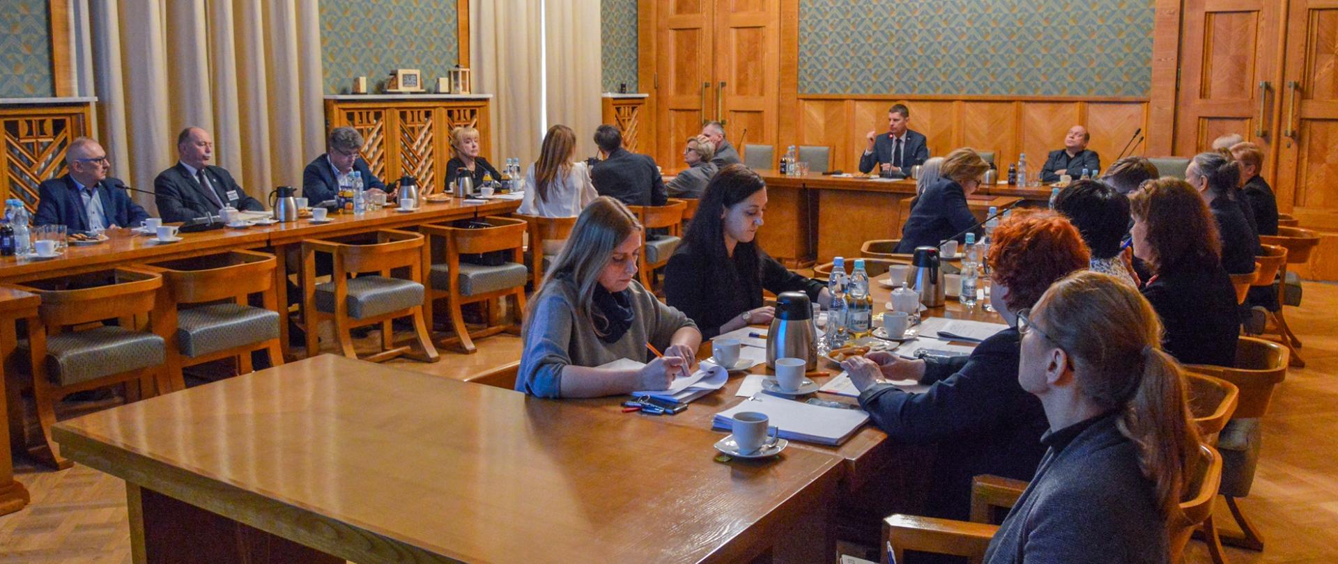 Widok na salę z zielonymi ścianami wyłożonymi drewnianymi panelami, przy ustawionych w czworokąt stołach siedzą ludzie.