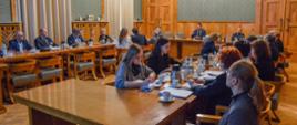 Widok na salę z zielonymi ścianami wyłożonymi drewnianymi panelami, przy ustawionych w czworokąt stołach siedzą ludzie.