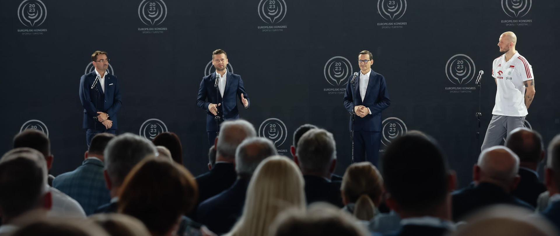 Minister Kamil Bortniczuk przemawia do gości ze sceny. Na scenie stoją także trzej inni mężczyźni - prezes Polskiej Siatkówki Sebastian Świderski, premier RP Mateusz Morawiecki oraz kapitan siatkarskiej kadry Bartosz Kurek.