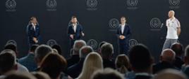 Minister Kamil Bortniczuk przemawia do mikrofonu na scenie podczas ogłoszenia decyzji o tym, że Polska zorganizuje siatkarskie mistrzostwa świata w 2027 roku. Na scenie po lewej stronie od ministra stoi prezes Sebastian Świderski z Polskiej Siatkówki. Po prawej stoi premier Mateusz Morawiecki. Obok premiera Bartosz Kurek, kapitan polskich siatkarzy.