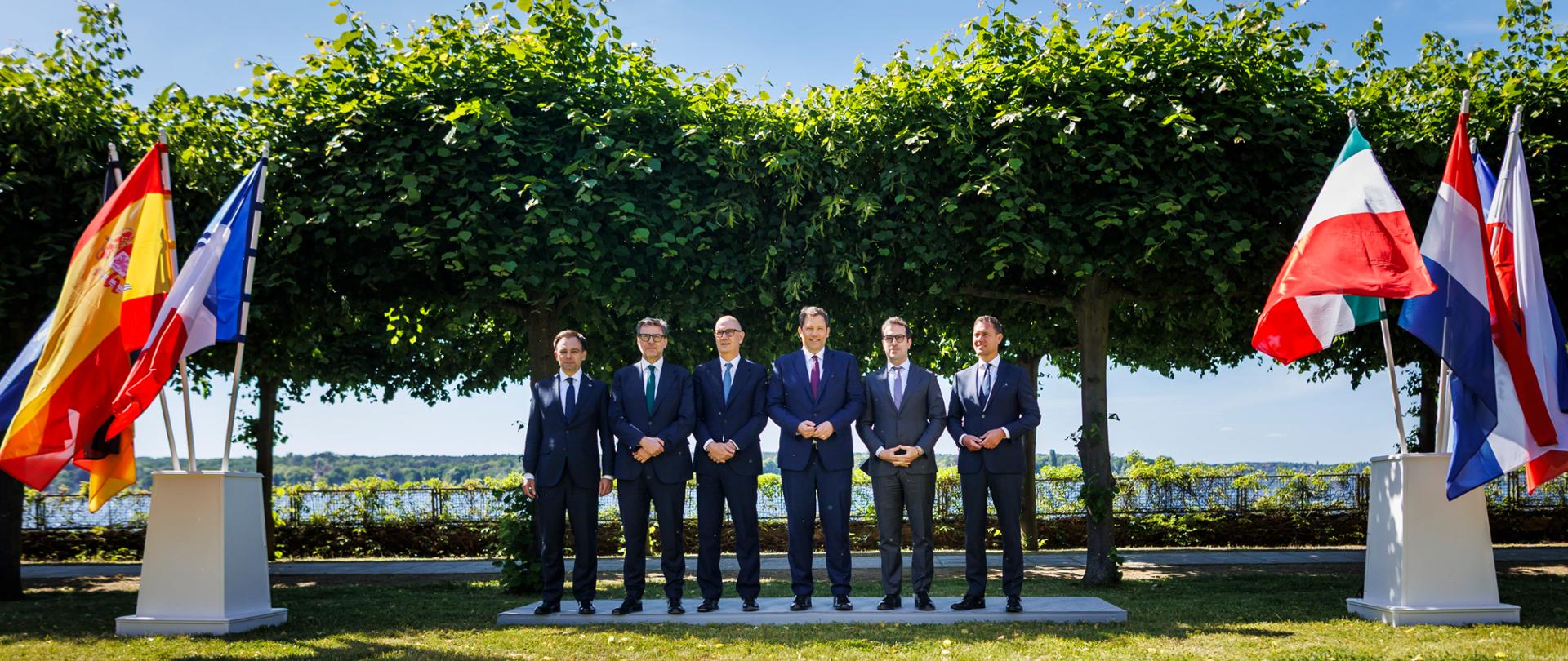 Andrzej Domański na wspólnym zdjęciu z ministrami finansów Francji, Niemiec, Włoch, Holandii i Hiszpanii. Po obu stronach ustawione są stojaki z flagami kilku państw europejskich, m.in. Polski, Włoch, Francji, Niemiec i Hiszpanii. W tle widać rząd drzew formujących zielony baldachim oraz jezioro. Osoby są ubrane w formalne stroje i stoją przodem do fotografującego, w słoneczny dzień.