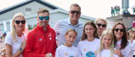 Kilkoro dzieci w sportowych strojach stoi na dworze, obok nich mężczyzna w czerwonym dresie, mężczyzna w białej koszuli i kobieta w białej koszulce.