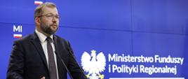 Minister funduszy i polityki regionalnej Grzegorza Puda stoi przy mównicy i przemawia do mikrofonów, za jego plecami ekran informujący o finansowaniu budowy nowego toru podejściowego w porcie Świnoujście