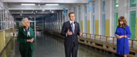 Na szkolnym korytarzu na błyszczącej podłodze stoją trzy osoby przy trzech mikrofonach - wiceminister Machałek, premier Morawiecki i kobieta ubrana na niebiesko.