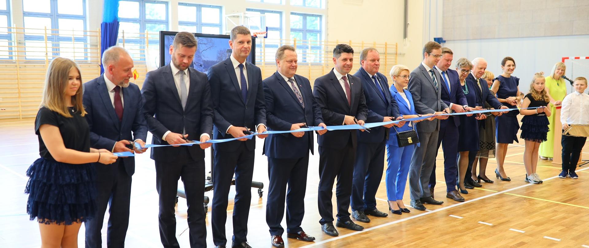 Uczestnicy wizyty stoją w rzędzie na sali gimnastycznej i trzymają niebieską wstęgę.