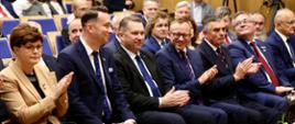 Widok z przodu pod kątem na ludzi siedzących na sali, w pierwszym rzędzie minister Czarnek, kobieta w żółtej marynarce i kilku mężczyzn w garniturach.
