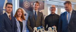 Minister i czworo młodych ludzi oglądają stojące na przykrytym niebieskim materiałem urządzenie - niebieskie rury w białym sześciokątnym szkielecie.