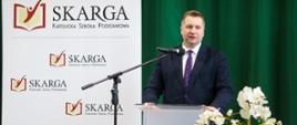 Minister Czarnek stoi za mównicą i mówi do mikrofonu, obok kwitnąca na biało roślina, za nim ścianka z napisem Skarga, w tle ciemnozielona kotara.