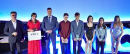 W oświetlonej na niebiesko sali wiceminister Piontkowski stoi wśród grupy młodych ludzi, obok niego kobieta w czerwonej sukience trzyma kartkę z napisem 6 000 zł.