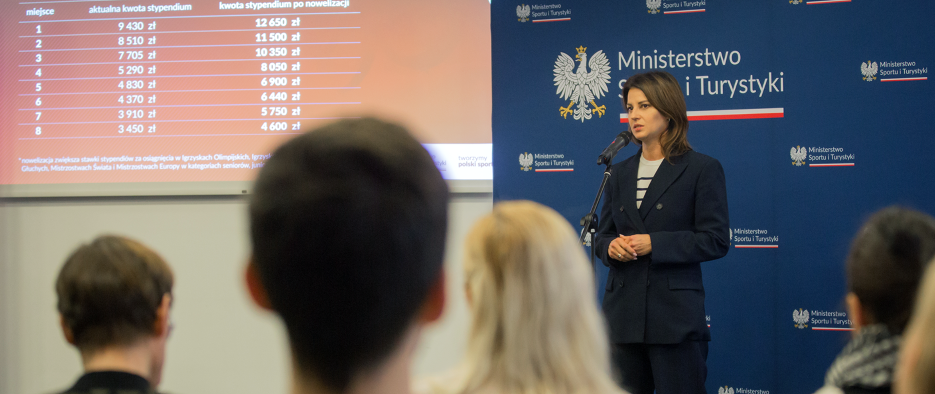 Minister Danuta Dmowska-Andrzejuk podczas briefingu dotyczącego zwiększonych stawek stypendiów dla sportowców - pani minister stoi na tle ścianki z logotypami ministerstwa, na ekranie obok wyświetlane są przykładowe stawki stypendiów