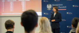 Minister Danuta Dmowska-Andrzejuk podczas briefingu dotyczącego zwiększonych stawek stypendiów dla sportowców - pani minister stoi na tle ścianki z logotypami ministerstwa, na ekranie obok wyświetlane są przykładowe stawki stypendiów