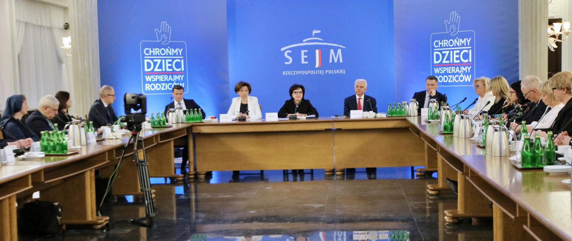 Mężczyźni i kobiety siedzą za stołami ustawionymi w literę U. Na stole mikrofony oraz butelki z wodą i czajniki.