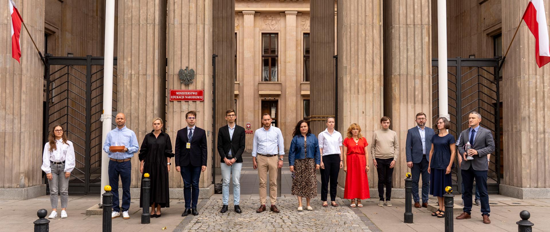 Zdjęcie przedstawia Minister Edukacji i pracowników ministerstwa przed budynkiem, oddających cześć podczas minuty ciszy