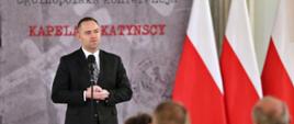Na tle ścianki z napisem Kapelani katyńscy stoi mężczyzna w czarnym garniturze i mówi do mikrofonu na stojaku, z boku trzy polskie flagi.