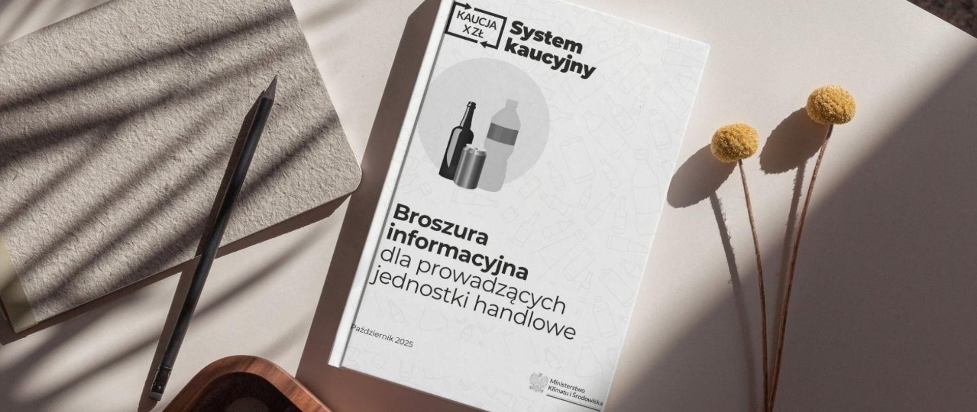 Na zdjęciu jest widoczna książka leżąca na stole. Na niej widnieje napis w lewym górnym rogu: System kaucyjny. Obok niego znajduje się logotyp Systemu kaucyjnego. Poniżej znajduje się napis: Broszura informacyjna dla prowadzących jednostki handlowe, październik 2025. W prawym dolnym rogu znajduje się logotyp Ministerstwa Klimatu i Środowiska. Na stole leży również ołówek, kartka oraz kwiaty.