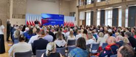 Widok z tyłu na wypełnioną siedzącymi na krzesłach ludźmi salę.