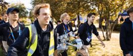 Czterech młodych ludzi idzie po trawie pod drzewami, niosą czterokołowego robota.