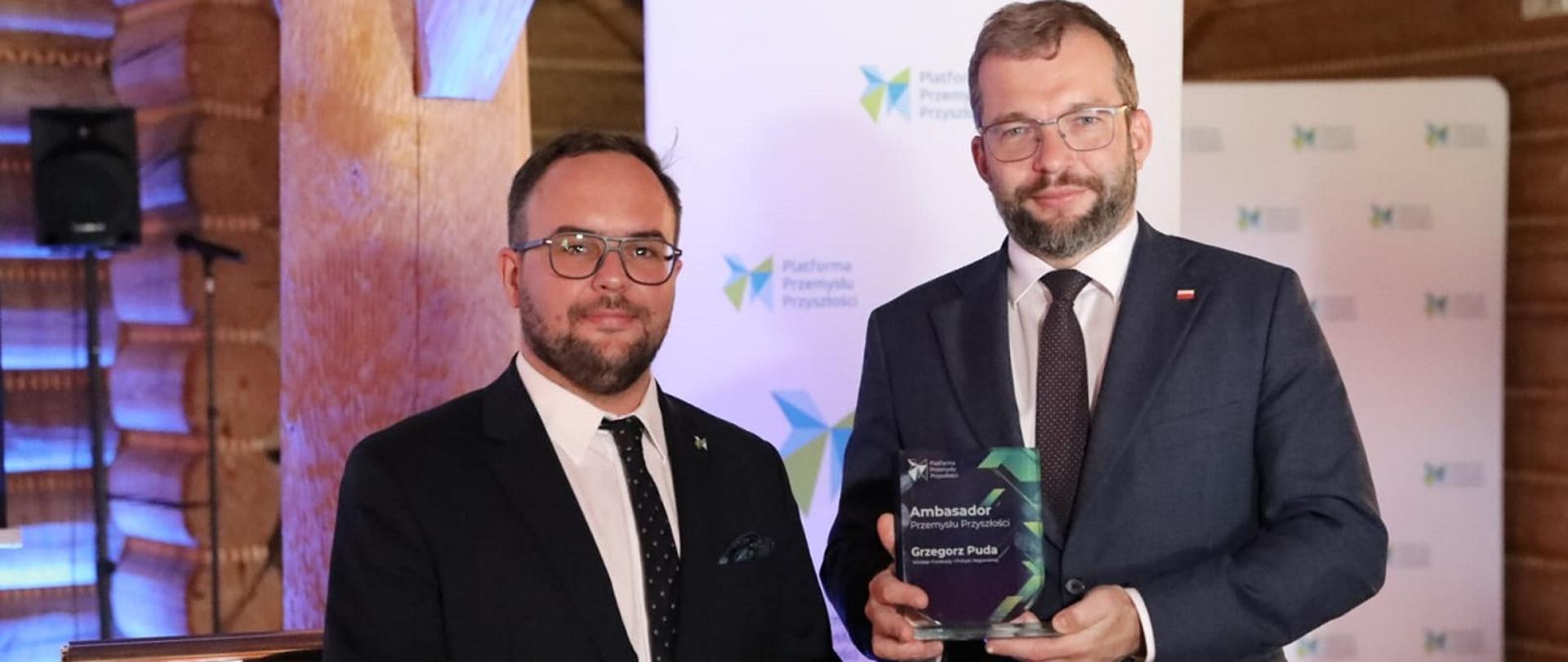 Minister Funduszy i polityki regionalnej Grzegorz Puda ze statuetką Ambasadora Przemysłu Przyszłości, minister trzyma w rękach statuetkę, obok niego stoi prezes zarządu Platformy Przemysłu Przyszłości Maciej Malik