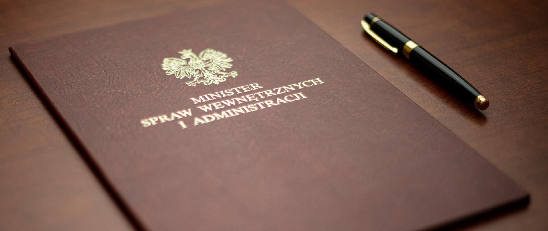 Na zdjęciu widać twardą, elegancką okładkę na dokumenty Ministra SWiA. Obok teczki leży eleganckie pióro do pisania. 
